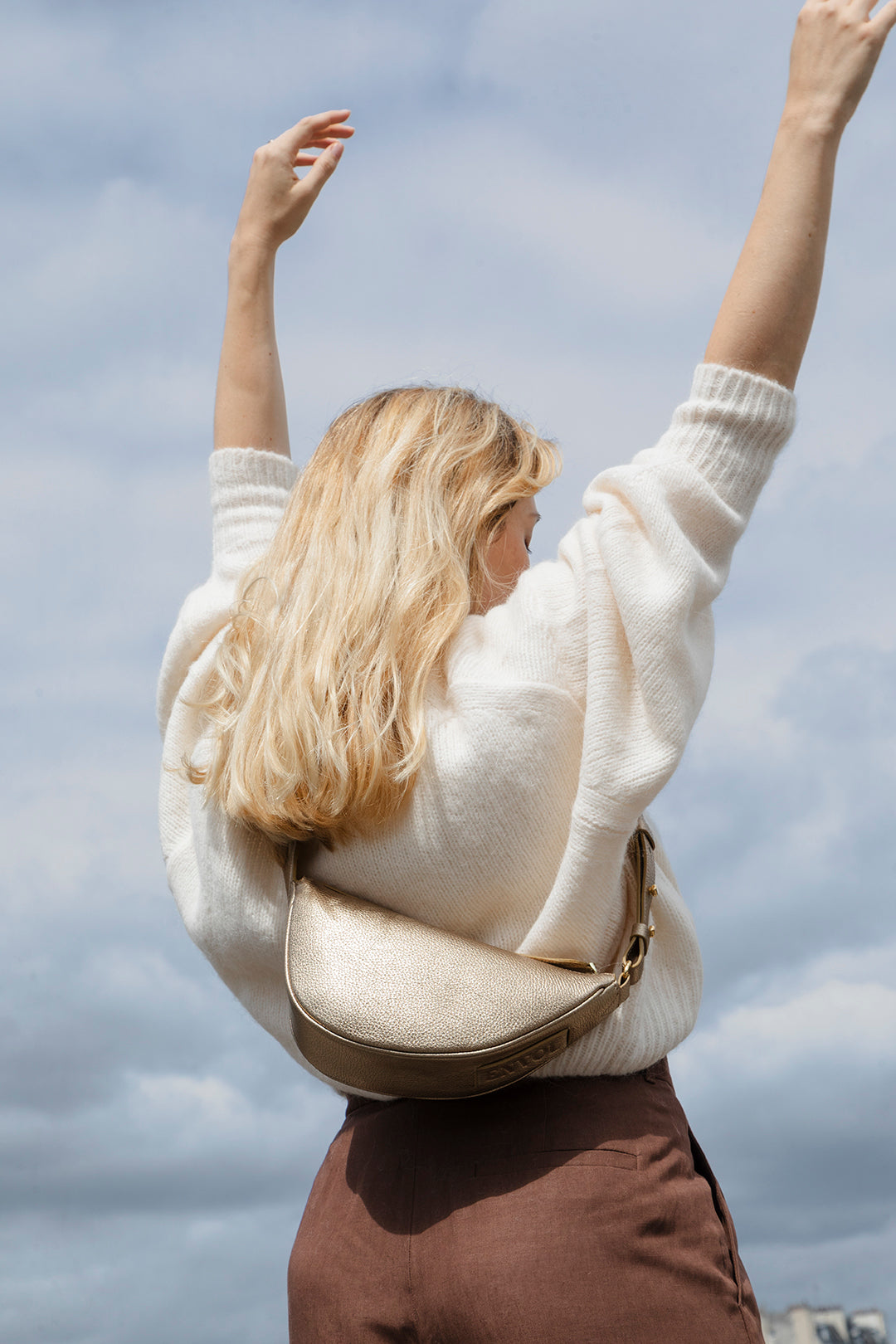 Femme portant le mini sac banane Lou ENVOL doré sous un ciel bleu #Couleur_Gold