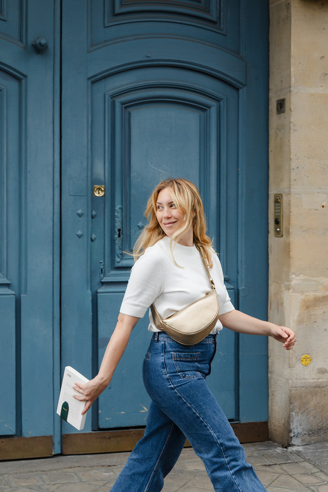 Femme portant le mini sac banane Lou ENVOL doré devant une porte bleue #Couleur_Gold