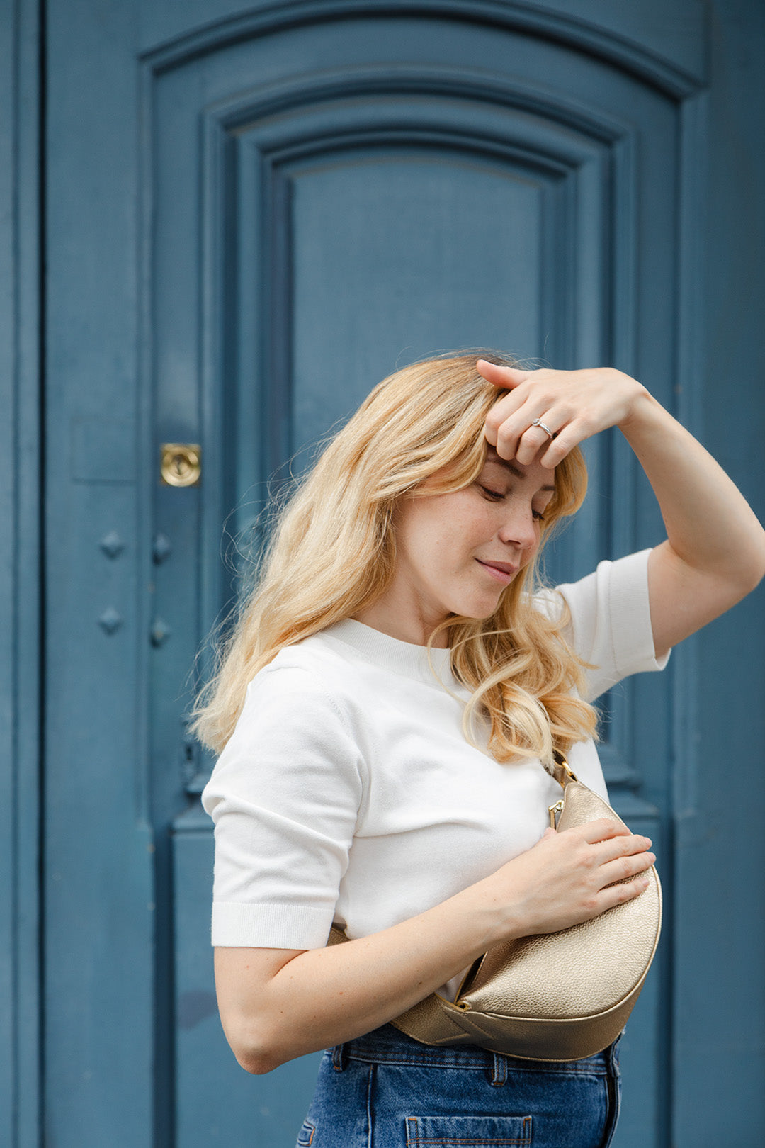 Femme portant le mini sac banane Lou ENVOL doré devant une porte bleue, de face #Couleur_Gold